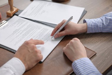 Belgeye işaret eden noter ve müşteri, kapalı oturulan ahşap masaya imza atıyor, yakın plan