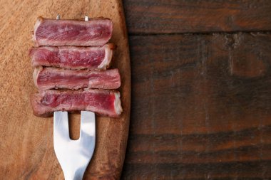 Carving fork with pieces of delicious beef on wooden table, top view. Space for text