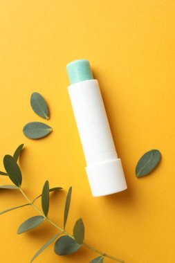 Dudak kremi ve okaliptüs yaprakları sarı arka planda, düz yerde.