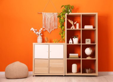 Wooden shelving unit with different home decor, houseplant and pouf near orange wall indoors