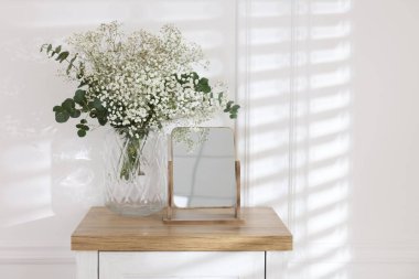 Beautiful gypsophila flowers with eucalyptus branches in vase and mirror on table at home. Space for text