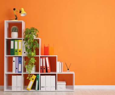 Shelving unit with folders and plants near orange wall indoors. Space for text