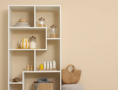 Shelving unit with dishware, jars of different products and other stuff near beige wall indoors, space for text
