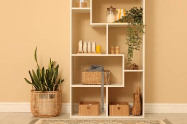 Shelving unit with dishware, jars of different products and other stuff near beige wall indoors