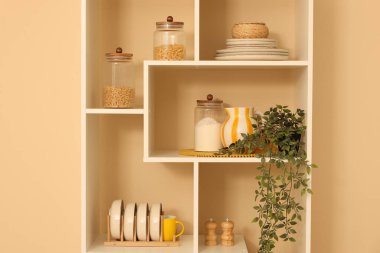 Shelving unit with dishware, jars of different products and other stuff near beige wall indoors