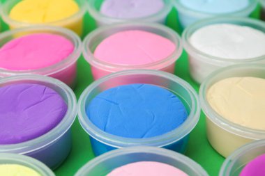 Different colorful modeling clay in plastic jars on green background, closeup