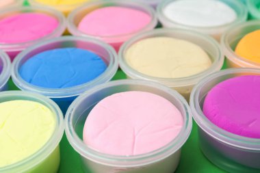 Different colorful modeling clay in plastic jars on green background, closeup