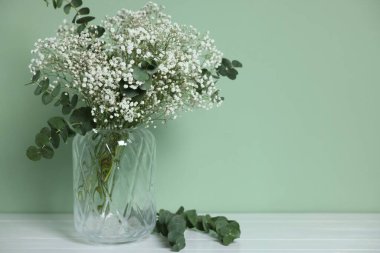 Beautiful gypsophila flowers and eucalyptus branches in vase on white table against turquoise background. Space for text
