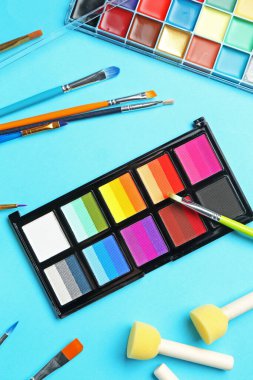 Açık mavi arka planda parlak yüz boyaları, fırçalar ve süngerler, düz yüz.