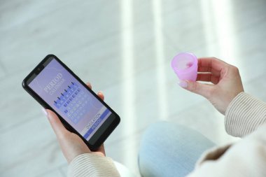 Regl. Adet fincanı kullanan bir kadın kapalı alanda akıllı telefondan takvim uygulaması kullanıyor.