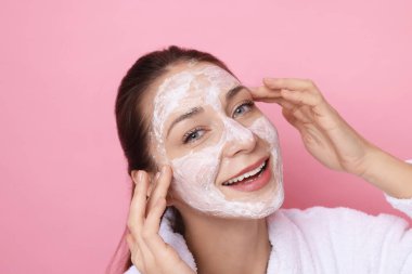 Spa günü. Güzel bir kadın pembe arka planda yüzüne maske takıyor.