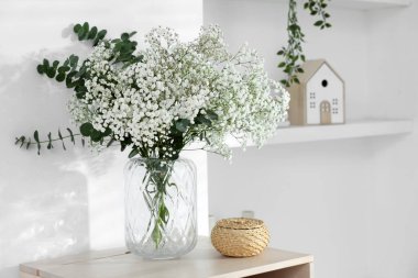 Bouquet of beautiful gypsophila flowers and eucalyptus branches in vase on chest of drawers near white wall