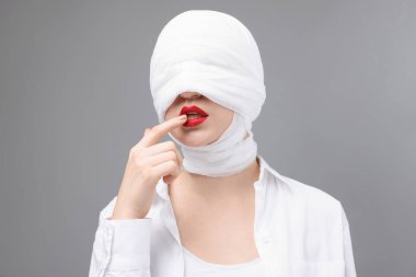 Gri arkaplandaki estetik ameliyattan sonra yüzü tıbbi bandaja sarılı bir kadın.