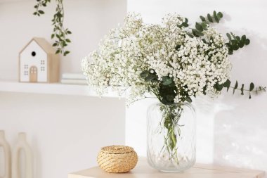 Bouquet of beautiful gypsophila flowers and eucalyptus branches in vase on chest of drawers near white wall