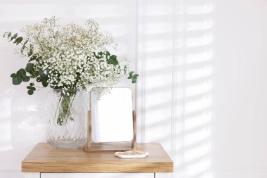 Beautiful gypsophila flowers with eucalyptus branches in vase and mirror on table at home. Space for text