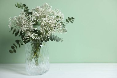 Beautiful gypsophila flowers and eucalyptus branches in vase on white table against turquoise background. Space for text