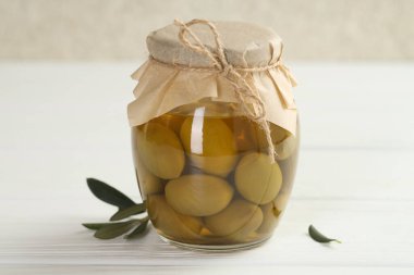 Cam kavanozda yeşil zeytin turşusu ve beyaz ahşap masada yapraklar, yakın plan.