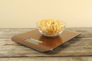 Elektronik mutfak terazisi, ahşap masada bej arka planda bir kase çiğ makarna, yakın plan.