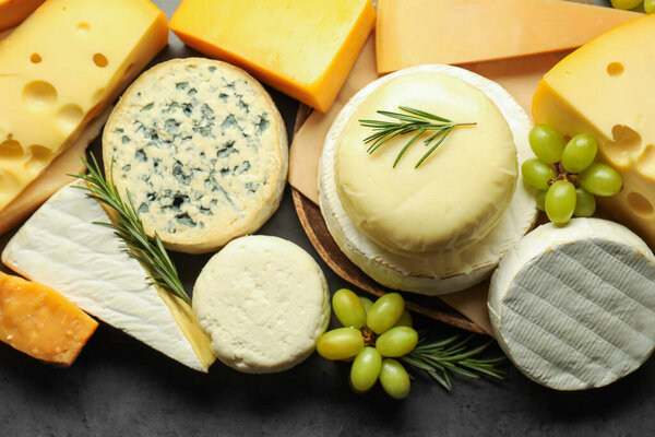Different types of cheese, rosemary and grapes on dark textured table, flat lay