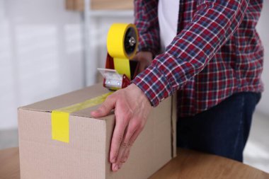 Karton kutuyu kapalı bir yere kapatmak için yapıştırıcılı bant kullanan bir adam.