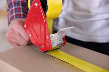 Karton kutuyu kapalı bir yere kapatmak için yapıştırıcılı bant kullanan bir adam.