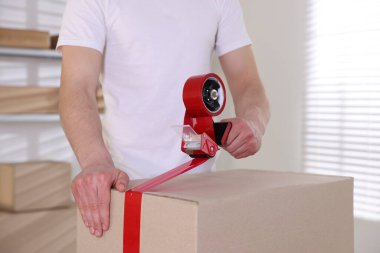 Karton kutuyu kapalı bir yere kapatmak için yapıştırıcılı bant kullanan bir adam.