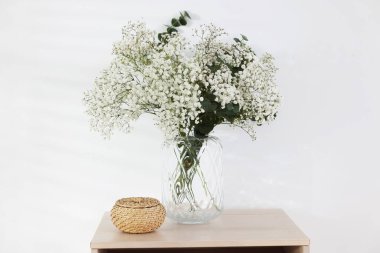 Bouquet of beautiful gypsophila flowers and eucalyptus branches in vase on chest of drawers near white wall