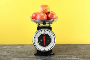 Mekanik mutfak terazisi, sarı arka planda koyu dokuma masa üzerinde elma kasesi.