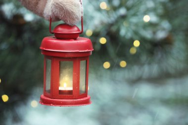 Woman holding Christmas lantern with burning candle in snowy park, closeup and space for text. Bokeh effect