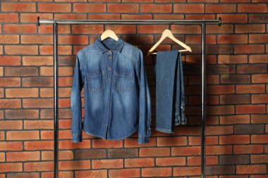 Stylish denim jacket and jeans hanging from rack near brick wall