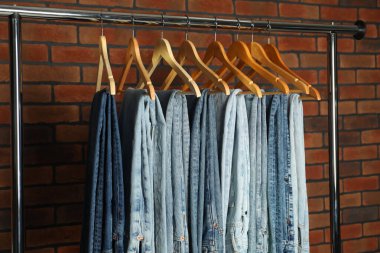 Many stylish jeans hanging from rack near brick wall