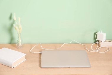 Kablolu USB adaptörü elektrik kablosuna takılı, yeşil duvarın yanındaki ahşap masada dizüstü bilgisayarı şarj ediyor.