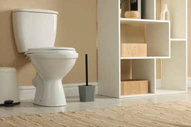 Toilet bowl, brush and trash bin near beige wall in restroom