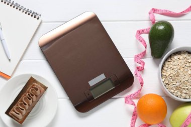 Elektronik mutfak ölçeği, mezura, not defteri ve beyaz ahşap masa üzerindeki ürünler, düz zemin. Kalori kontrolü