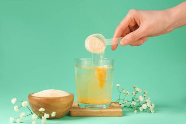 Kadın bardağa kolajen tozu ekliyor. Turkuaz arka planda su var. Yakın plan.