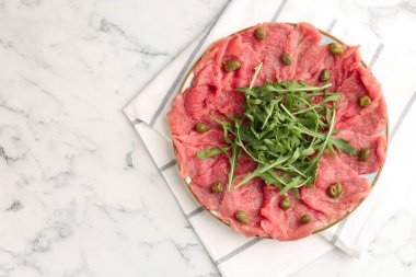 Delicious beef carpaccio with arugula and capers on white marble table, top view. Space for text