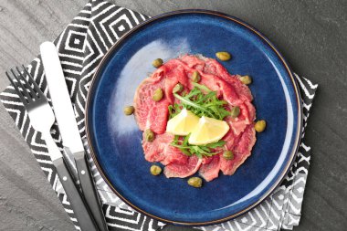 Fresh beef carpaccio with arugula, lemon and capers on dark textured table, top view