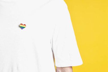 Sarı arkaplanda LGBT bayrağı renginde kalp şeklinde iğnesi olan adam, yakın plan