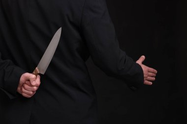 Sırtında bıçak tutan bir iş adamı siyah arka planda el sıkışma teklif ediyor.