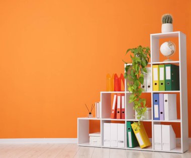 Shelving unit with folders and plants near orange wall indoors. Space for text