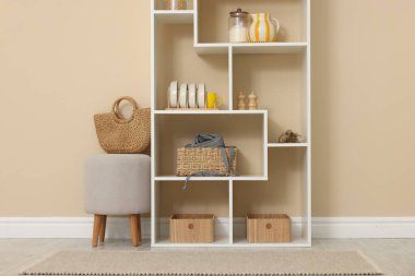 Shelving unit with dishware, jars of different products and other stuff near beige wall indoors