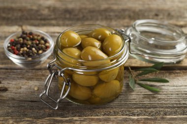 Cam kavanozda turşulu yeşil zeytin ve ahşap masada baharatlar, yakın plan.