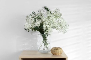 Bouquet of beautiful gypsophila flowers and eucalyptus branches in vase on chest of drawers near white wall
