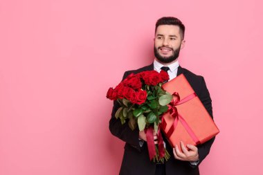 Kırmızı gül buketi ve pembe arka planda hediye kutusu olan mutlu bir adam.