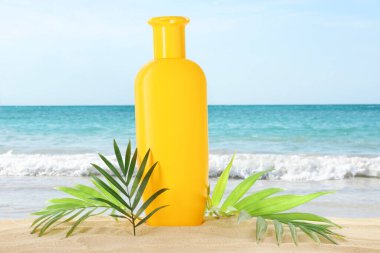 Bir şişe güneş kremi ve okyanus yakınlarındaki kumlu sahilde yeşil tropik yapraklar.