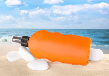 Deniz kenarındaki kumlu sahilde bir şişe güneş kremi ve taşlar.