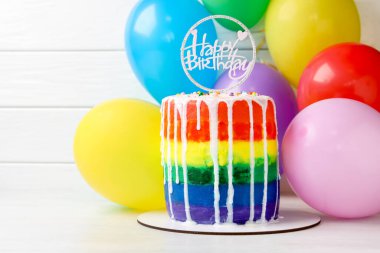 Lezzetli gökkuşağı doğum günü pastası. Masada beyaz ahşap arka planda balonlar var.
