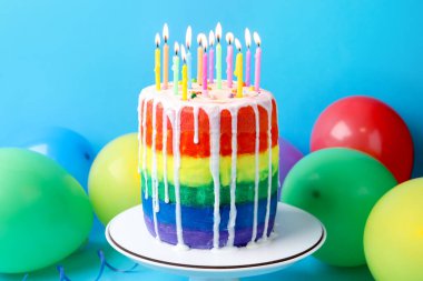 Enfes gökkuşağı pastası yanan mumlar ve açık mavi arka planda balonlar, yakın plan.