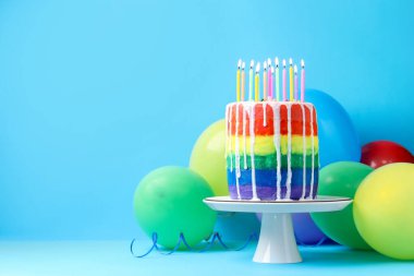 Lezzetli gökkuşağı doğum günü pastası yanan mumlar ve açık mavi arka planda balonlar, metin için alan