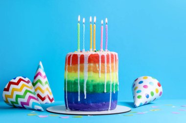 Enfes gökkuşağı doğum günü pastası yanan mumlar, parti şapkaları ve açık mavi arka planda konfeti